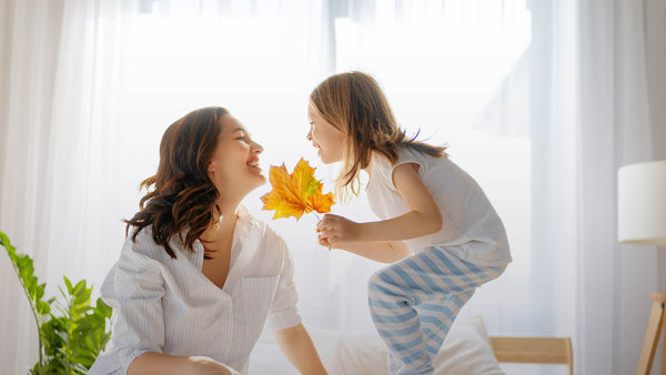 image of mother and daughter on a bed smiling at each other; daughter is in pajamas holding fall leaves but it's daylight outside. Image is used in a blog post about fall season change and taking melatonin for sleep.